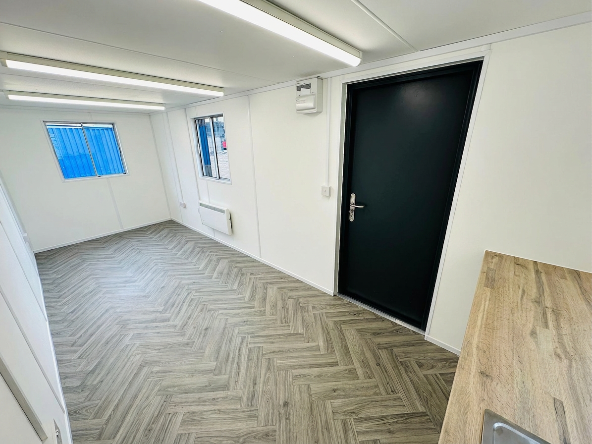 Refurbished anti-vandal office container interior with herringbone vinyl flooring, white walls, and a black steel door