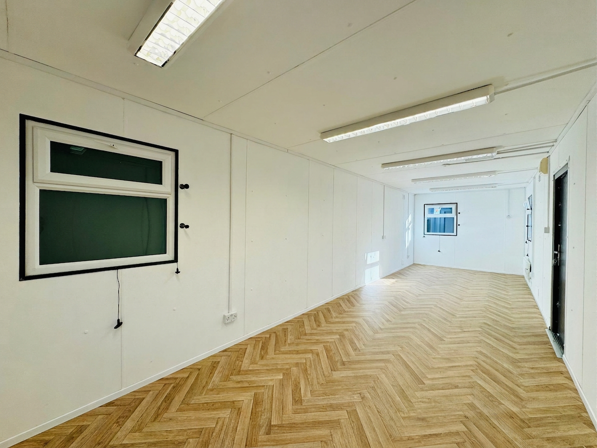 Refurbished 32ft shipping container office interior with white walls, herringbone wood floors, and fluorescent lights