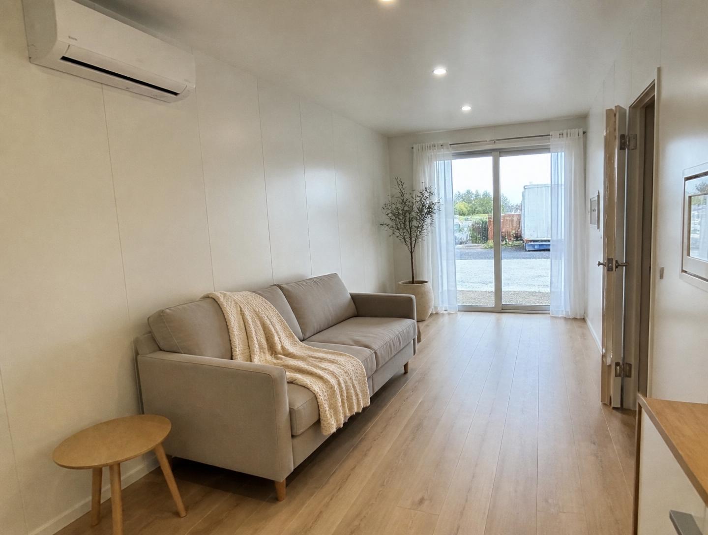 Luxury 40ft prefab container house interior — living area reverse angle with linen sofa, sheer curtains, and mini-split HVAC